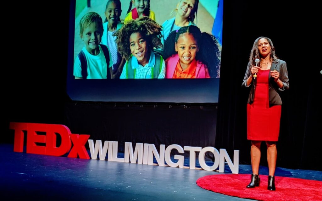 A Black woman wearing a red dress and gray blazer stands on stage as she gives a TEDx talk.